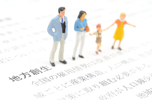 「地方創生」について書かれたテキストの上に立つミニチュアの家族