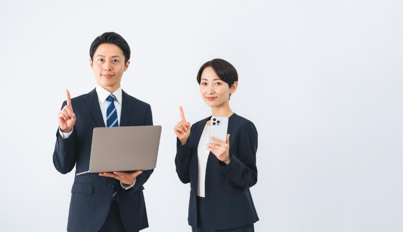 それぞれPCとスマホを片手に持って人差し指を立てる男女